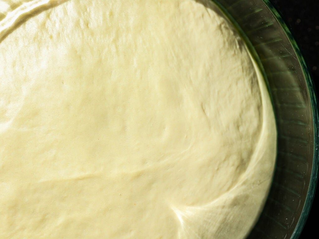 A close-up of smooth, risen dough in a glass bowl, ready for baking. The surface is slightly glossy, with an even texture, illuminated by soft light.